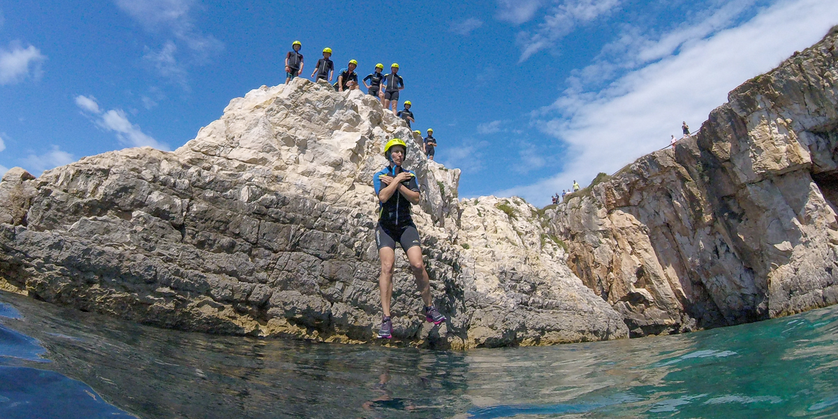 Groups of people standing on the cliffs while one woman is jumping in the sea.