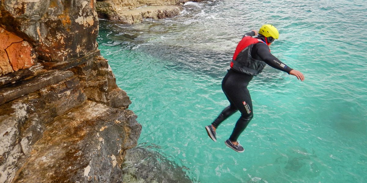 A man equipped with wetsuit, life vest and a helmet jumping off the rocks into the sea.
